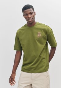 Olijfgroen katoenen T-shirt met korte mouwen en een ronde halslijn. Heeft een bedrukt tekst "Milan. Parijs. Berlijn." aan de linkerkant.