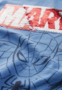 Sininen puuvillapaita, jossa on paljettikoristeinen "MARVEL"-teksti punaisella ja valkoisella, ja musta Spider-Man -kuvio tulostettuna.