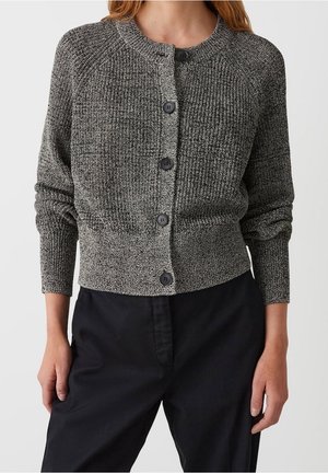 Femme portant un cardigan gris texturé à boutons et un pantalon noir, debout, les mains détendues le long du corps, devant un fond uni.