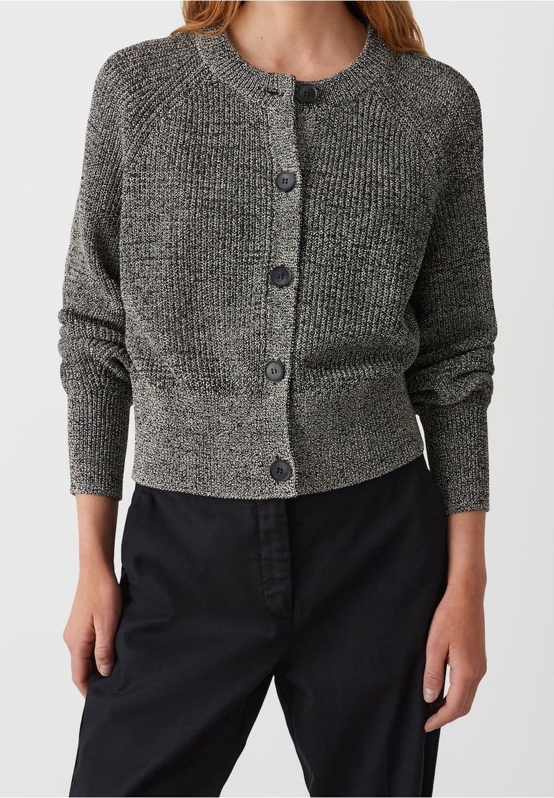 Femme portant un cardigan gris texturé à boutons et un pantalon noir, debout, les mains détendues le long du corps, devant un fond uni.