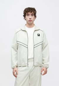 Ελαφρύ, κρεμ μπουφάν με μαύρη επένδυση και μικρό έμβλημα. Διαθέτει φερμουάρ και ελαστικές μανσέτες, συνδυασμένο με ταιριαστό σύνολο.