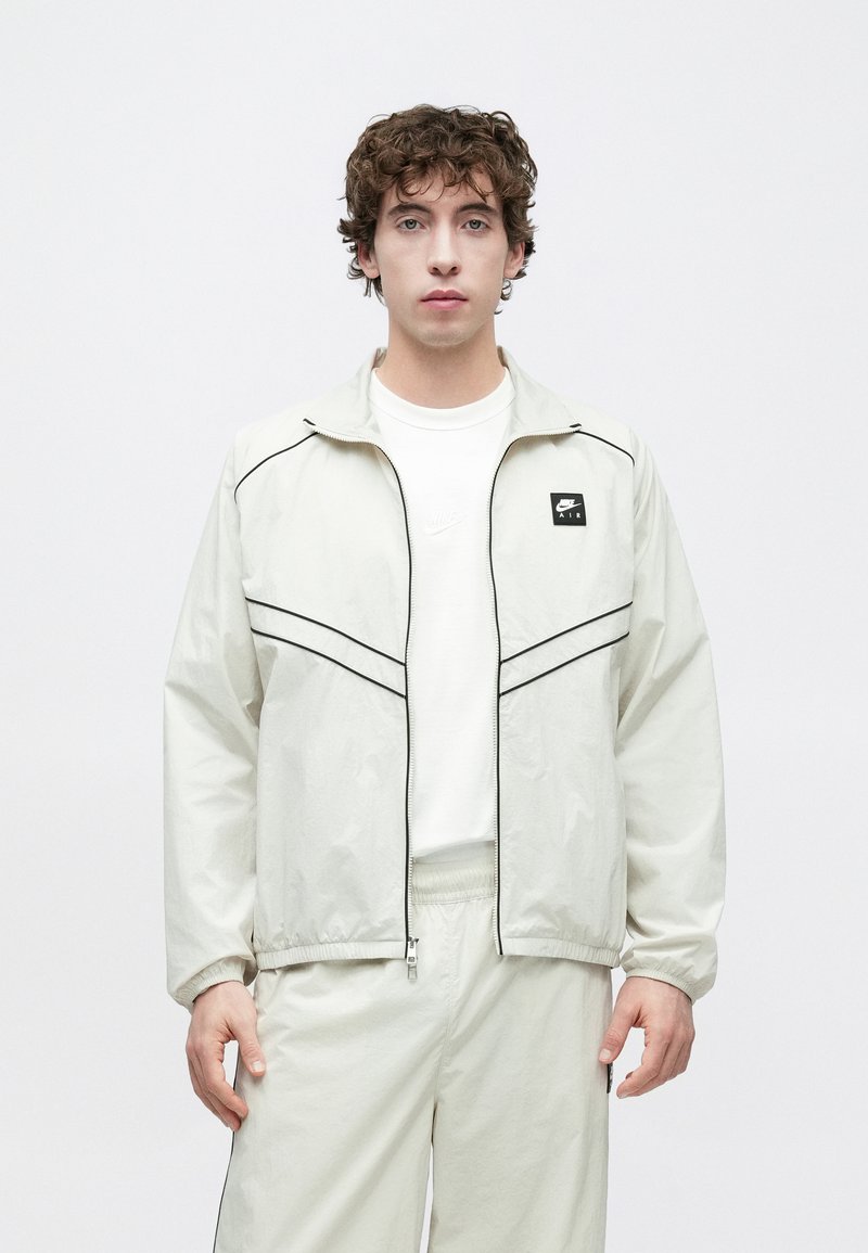 Chaqueta ligera de color crema con ribete negro y un pequeño parche con logo. Cuenta con cierre de cremallera y puños elásticos, combinada con un conjunto a juego.