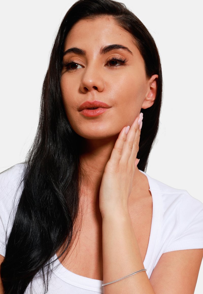 Modella con lunghi capelli neri e trucco neutro, indossa una maglietta bianca, toccandosi delicatamente il viso, mostrando un sottile braccialetto d'argento al polso.