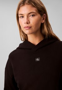Ung kvinde med lysebrunt hår iført en sort hættetrøje med et lille Calvin Klein-logo på brystet mod en neutral baggrund.