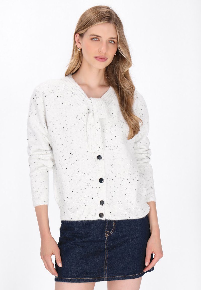 Hvidprikket strikcardigan med slips ved halsen, med knaplukning og lange ærmer. Bæret over en mørk denimnederdel.