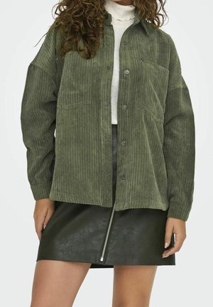Kvinde iført en oversized olivengrøn corduroy-jakke over en hvid polotrøje og en mørkegrøn læder mini-nederdel med lynlås foran.