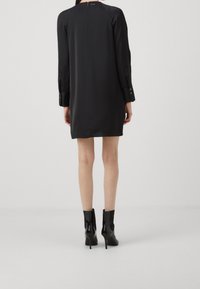 Frau, die ein lockeres schwarzes Kleid mit langen Ärmeln und schwarze, glänzende, hochhackige Stiefeletten trägt, von hinten gezeigt.