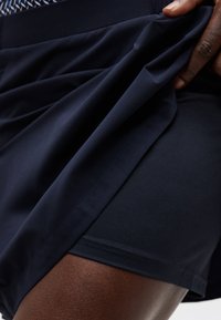 Χέρι σηκώνει το εξωτερικό στρώμα σορτς για να αποκαλύψει εφαρμοστά εσωτερικά κοντά σορτς συμπίεσης σε ναυτικό μπλε πάνω σε μηρό με σκούρο τόνο δέρματος.