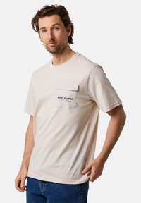 Ljusbeige t-shirt med kort ärm, tillverkad av bomull, med en framfick med lock och tryckt logotyp. Enkel, avslappnad passform.