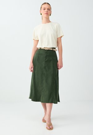 Femme marchant, portant un haut crème à manches courtes, une jupe midi verte avec une ceinture beige, des boucles d'oreilles vertes et des sandales marron sur fond blanc.