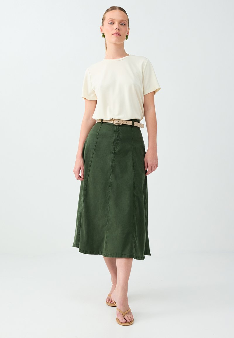 Femme marchant, portant un haut crème à manches courtes, une jupe midi verte avec une ceinture beige, des boucles d'oreilles vertes et des sandales marron sur fond blanc.