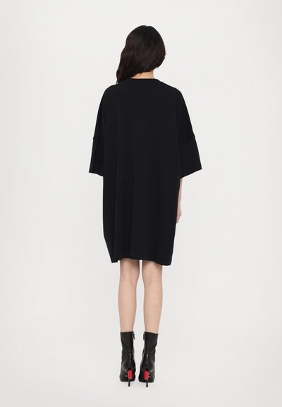 Μαύρο oversize φόρεμα με κοντό μανίκι, χαλαρή εφαρμογή και απαλό υλικό, συνδυασμένο με ψηλές μαύρες μπότες με κόκκινες λεπτομέρειες.