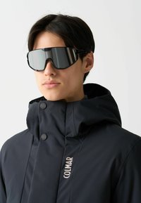 Svart isolerad jacka med hög krage, tryckknappar och en framträdande logotyp. Modellen bär släta, spegelbelagda solglasögon som visar upp en sportig design.