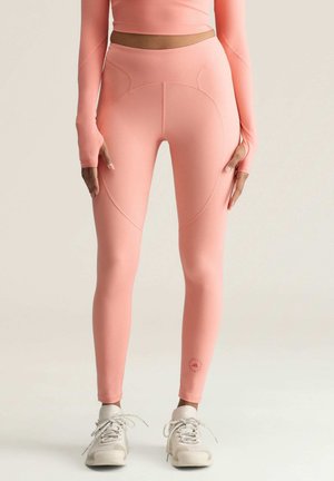 Person trägt korallenpinke Leggings mit hohem Bund und passendes langärmeliges Croptop, kombiniert mit weißen Sneakern, steht vor hellem Hintergrund.