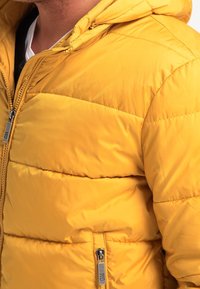 Homme portant une veste matelassée jaune avec des fermetures éclair visibles et une chemise blanche en dessous, montré du cou jusqu'au milieu du torse.