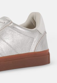 Metalické stříbrné tenisky s texturovaným povrchem, smetanovým límcem a hnědou gumovou podrážkou. Obsahují klasický design se šněrováním a prošívané detaily.