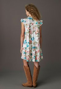 Robe fleurie avec des manches courtes à volants, fond blanc, fleurs colorées en rose et bleu, avec dentelle en finition et ourlet à volants. Portée avec des bottes marron.