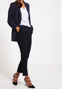 Žena ve tmavě modrém blejzru, bílém topu s výstřihem do V a odpovídajících tmavě modrých kalhotách, v černých sandálech na vysokém podpatku, stojí s rukama v kapsách.
