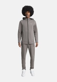 Grå atletisk sweatshirt og joggers i et glat stof. Har en hætte, lynlås foran og hvide logo-detaljer. Hvide sneakers fuldender outfittet.