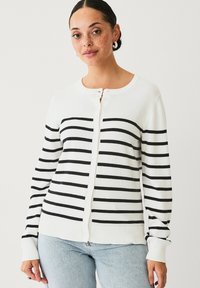 Cardigan bianco con strisce orizzontali nere, dotato di colletto rotondo, chiusura con bottoni e polsini e orlo a coste. Indossato con jeans blu.