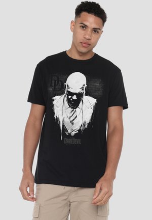 Young man wearing black t-shirt with white graphic of a serious bald man in suit and tie, standing against a plain light background.