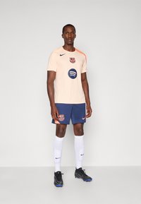 Licht perzikkleurig voetbalshirt met een oranje schouderaccent, Barcelona-logo, blauw Spotify-logo. Donkerblauwe shorts met logo, witte sokken, zwarte schoenen.