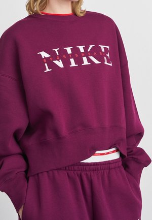 Personne portant un sweat-shirt de sport Nike violet et un pantalon assorti, les mains dans les poches, laissant apparaître une partie d'une ceinture blanche en dessous.