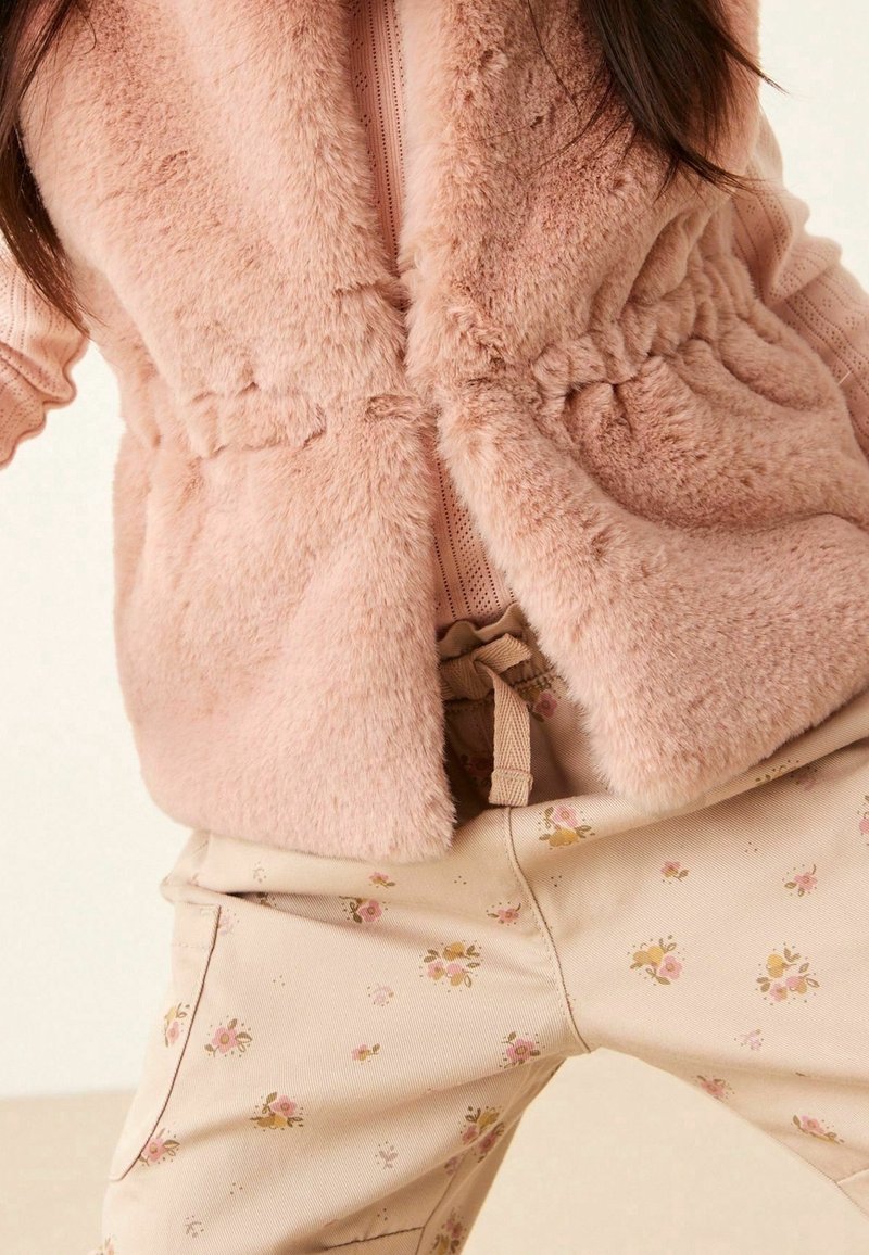 Gilet en fausse fourrure rose pâle avec détail de taille froncé, associé à un pantalon beige ornés de petits motifs floraux roses et dorés.