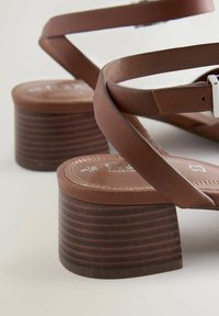 Hnedé kožené sandále s hrubými kvádrovými podpätkami a nastaviteľnými remienkami okolo členkov na svetlom povrchu.