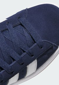 Primo piano di una sneaker in pelle scamosciata blu con tomaia strutturata, strisce bianche e lacci intrecciati. Suola in gomma visibile nella parte inferiore.