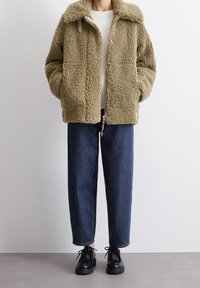 Luddig olivgrön jacka med mjuk textur, framdragkedja och tryckknappsstängning. Matchas med mörka denimbyxor och svarta snörskor.