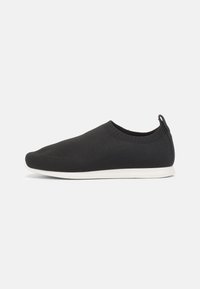 Svart strikket slip-on sko med en tett overdel, hvit gummisåle og en liten løftestropp på hælen for enkel påkledning.