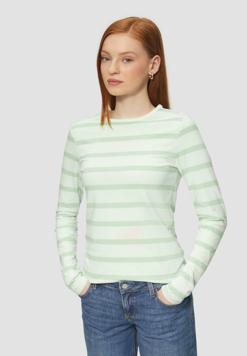Lange mouwen, lichtgroene gestreepte top met een ronde halslijn, gemaakt van zachte stof. Gecombineerd met blauwe jeans, handen in de zakken.
