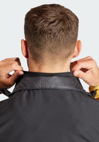 Schwarze Jacke mit Stehkragen, der Stoff hat eine glatte Textur. Die Hände passen den Kragen im Rücken an und zeigen ein mehrschichtiges Design.