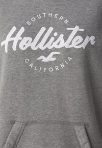 Sudadera gris con un logo bordado de "Hollister" y el texto "Southern California". Incluye un bolsillo frontal y una tela texturizada.