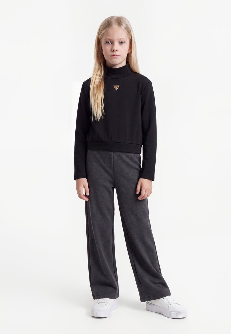 Jeune fille aux longs cheveux blonds portant un haut noir à manches longues, un pantalon gris large et des baskets blanches, debout sur un fond blanc.
