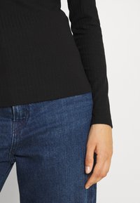 Maglietta nera a maniche lunghe con texture a coste abbinata a jeans in denim blu scuro, che mettono in mostra una silhouette aderente e dettagli di cucitura visibili.