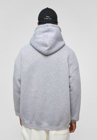 Grijze hoodie gemaakt van zacht materiaal, met een relaxte pasvorm, geribbelde manchetten en een kangoeroezak. De achterkant toont de detaillinie van de capuchon.