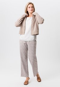 Hellbeige gestrickte Strickjacke mit langen Ärmeln, kombiniert mit einem weißen Oberteil und gemusterten, weit geschnittenen Hosen in Brauntönen. Braune Schluppen-Sandalen.