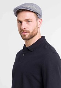 Pilkos spalvos plokščias kepurė iš tekstūruotos medžiagos, užsidėta su juodu polo marškinėliu, turinčiu apykaklę ir sagų detalę. Artimas portretinis vaizdas.