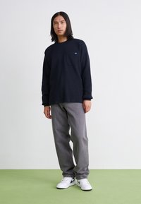 Μαύρη μπλούζα με μακριά μανίκια και τσέπη στο στήθος, σε συνδυασμό με γκρι παντελόνι και λευκά αθλητικά παπούτσια με μαύρο σήμα. Απλός, υφασμένος σχεδιασμός.