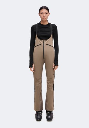 Personne portant un pantalon de neige beige à bretelles avec des fermetures éclair noires, un haut noir à manches longues et des chaussures de ski noires, se tenant droite devant un fond uni.