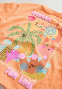 Narancssárga póló játékos trópusi grafikákkal, köztük mosolygó nappal és növényekkel, emelt rózsaszín "HAVANA" és "FUN TIME" felirattal.