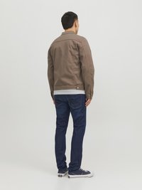 Brun bomulljakke med krage, kombinert med blå denimjeans og marineblå sneakers, sett fra bakgrunnen mot en hvit bakgrunn.