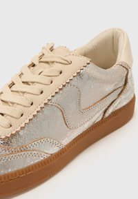 Sneaker features a beige leather upper with a cracked metallic sheen, textured accents, beige lace closure, and a rubber gum sole.