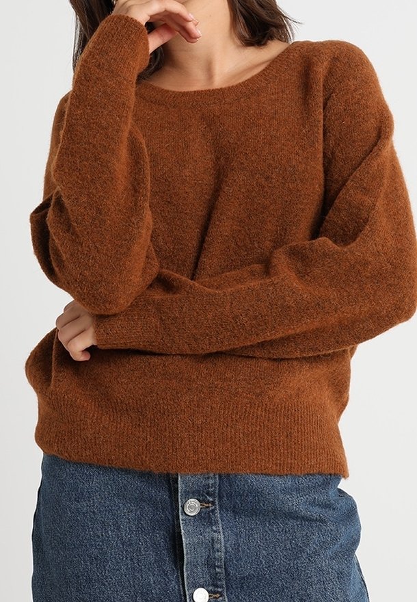Femme portant un pull en maille marron duveteux à manches longues et une jupe en jean bleue avec des boutons à l'avant, posant avec une main près du visage.
