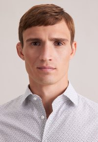 Camicia bianca con bottoni, decorata da un piccolo motivo ripetuto in blu scuro. Presenta un colletto classico e una texture di tessuto morbida.