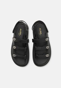 Svarta sandaler tillverkade av syntetiskt material med tre justerbara remmar med silverfärgad spänne, en texturerad fotbädd och en platt sula.