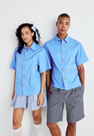 Two individuals wear light blue short-sleeve shirts with decorative buttons. One has a gingham skirt, the other striped shorts. Both have white tights.
