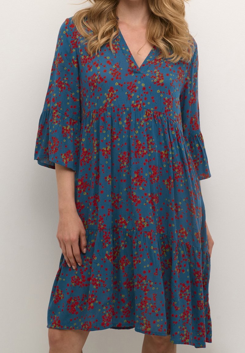 Femme portant une robe bleue longueur genou avec un motif floral rouge et jaune, col en V, et manches trois-quarts évasées.